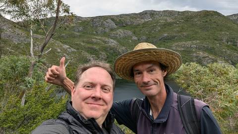      Two hikers taking a selfie with lake and rugged green hills in background.
  