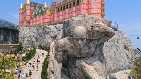       Visitors explore a theme park dominated by a colossal stone Titan statue and a colorful castle perched on rocky cliffs.
  