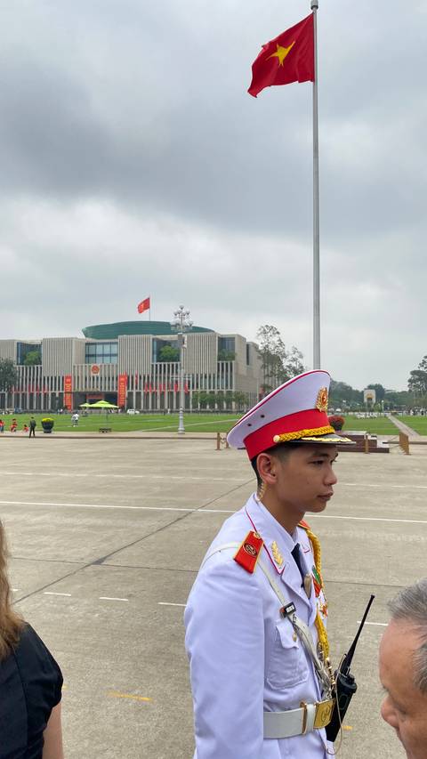       A uniformed guard with a red-and-white cap stands before the modern facade of a government building in Hanoi.
  