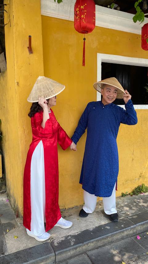       Two travelers in bright traditional Ao Dai and conical hats pose playfully against a vibrant yellow wall.
  