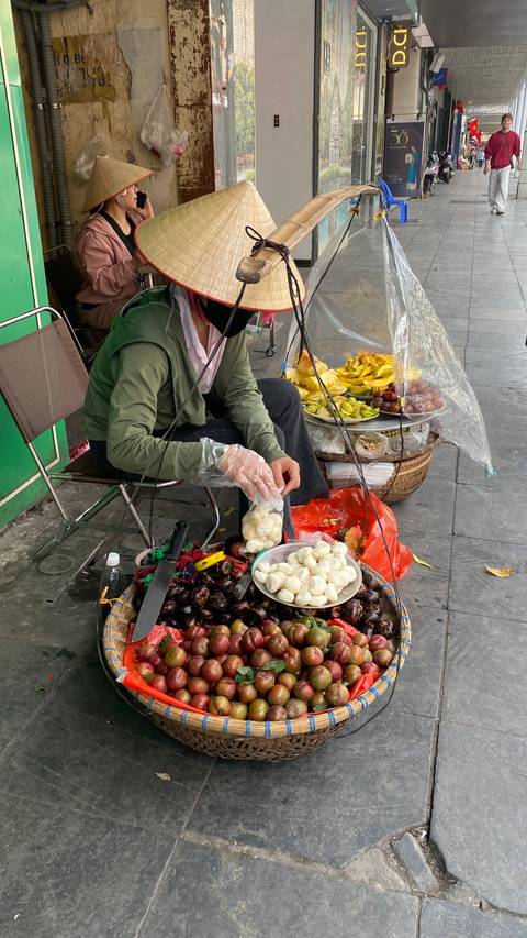       A street vendor wearing gloves packages tropical fruits at a small sidewalk stall.
  