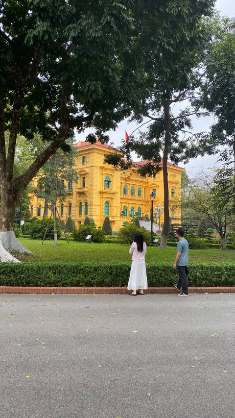       Two visitors admire the yellow French-colonial Presidential Palace surrounded by lush gardens in Hanoi.
  