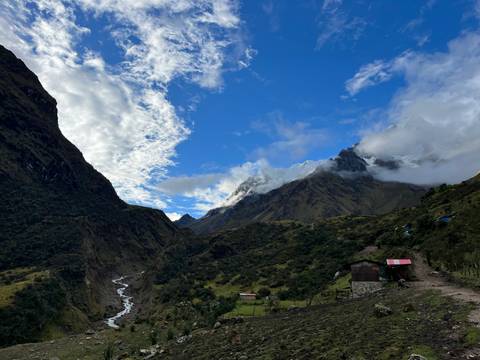       Sunny valley with steep green slopes, creek and rustic cabin beneath snowy mountain.
  