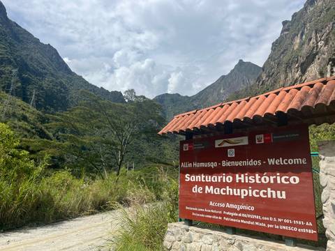       Entrance sign welcoming visitors to Machu Picchu sanctuary with lush peaks behind.
  