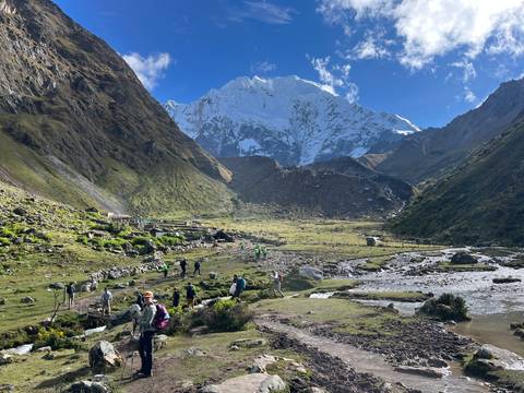       Trekkers crossing valley toward glacier-capped mountain, following stream through green pasture.
  
