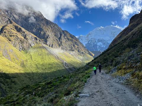       Backpackers walking along shadowed mountain trail toward snow-covered peak.
  