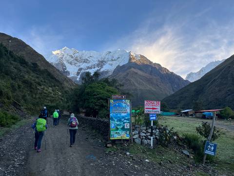      Trekkers with backpacks walk along a dirt road toward a dramatic snow-covered Andean peak under early morning light.
  