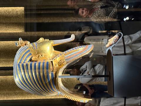       Side profile of Tutankhamun's iconic golden mask displayed behind a glass case with visitors reflecting.
  