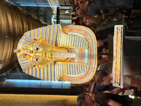       Front view of Tutankhamun's funerary mask surrounded by crowds inside a museum gallery.
  