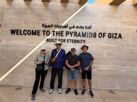       Four travelers pose beneath a sign welcoming visitors to the Pyramids of Giza.
  