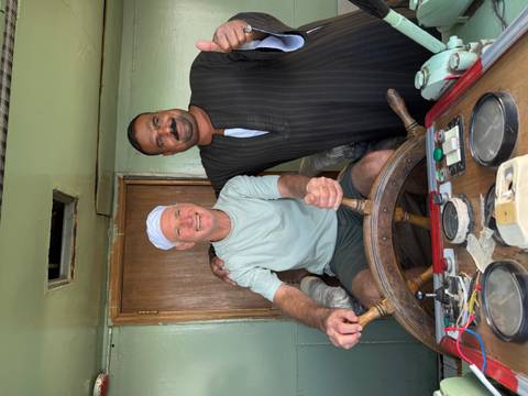       Two men, one steering a wooden ship's wheel, share a friendly moment aboard a riverboat.
  