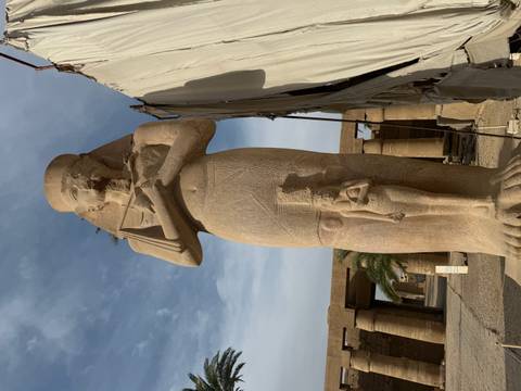       Tall sandstone pharaonic statue stands in open temple courtyard under hazy sky.
  