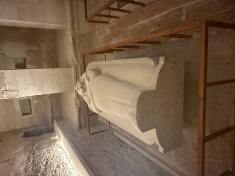       Stone sarcophagus in a dimly lit ancient Egyptian burial chamber with rough walls and protective railings.
  