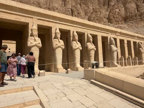       Colonnaded façade of the Temple of Hatshepsut with large Osiride statues and visitors walking across the forecourt.
  