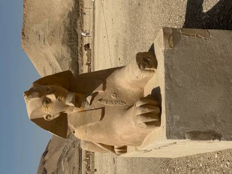       Sand-colored sphinx statue with human head and lion body positioned outdoors in a desert archaeological site.
  