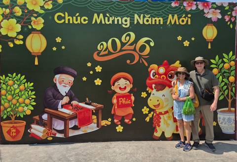       Smiling couple pose before a vibrant Tet New Year mural reading 'Chúc Mừng Năm Mới 2026'.
  