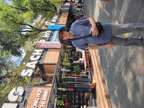       Tourist stands on a pedestrian book street lined with stalls under large 'Sách' signage and shady trees.
  