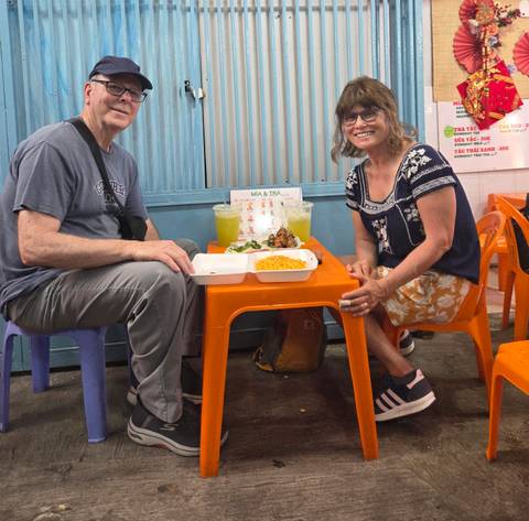       Two travellers sit on small plastic stools inside a casual eatery enjoying local Vietnamese street food.
  