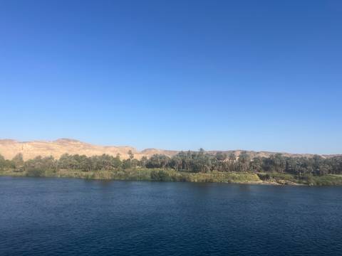       Blue waters of the Nile bordered by palm groves and desert hills under a cloudless sky.
  