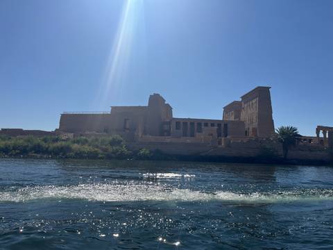       Ancient Philae Temple complex seen from a boat with bright sunlight creating a lens flare.
  