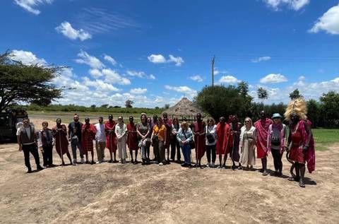       Large tour group standing with Maasai hosts in an open field beneath a bright blue Kenyan sky.
  
