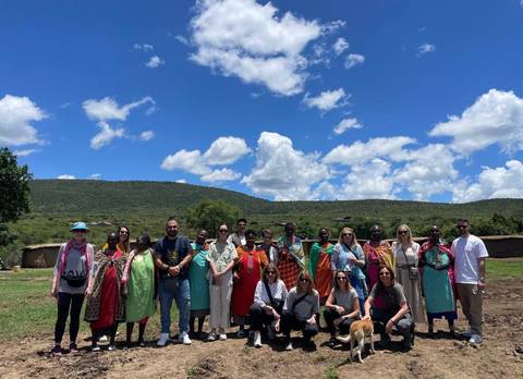       Travel group posing with Maasai women in front of green hills and traditional huts.
  
