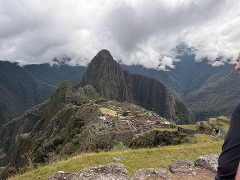       Iconic view of the Inca citadel of Machu Picchu surrounded by steep green mountains under a cloudy sky, with part of a visitor visible at the edge.
  