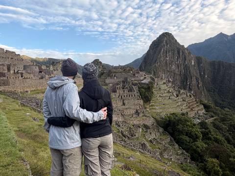       A couple dressed in trekking gear embrace while admiring the stone terraces of Machu Picchu at dawn.
  