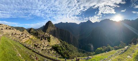       Sunlit panorama of Machu Picchu ruins with bright sky and dramatic clouds as the sun sets over the Andes.
  