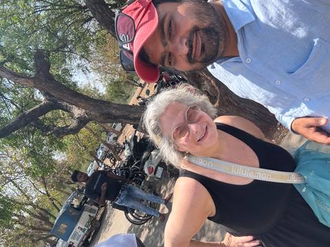       Selfie of traveler with guide under a leafy tree beside parked motorbikes on a hot day.
  