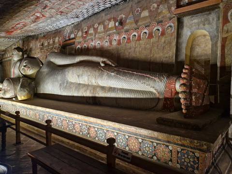       An ancient reclining Buddha statue lies within a richly painted cave temple hall.
  