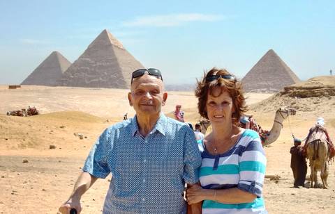       Two travellers posing with the Pyramids of Giza in the desert, camels and guides in the background.
  