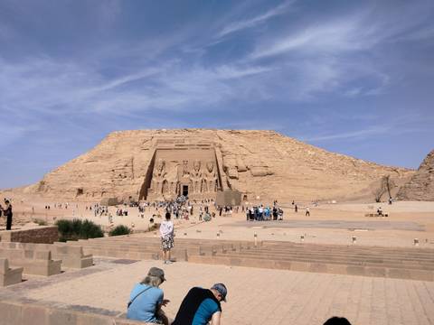       Wide view of the monumental façade of Abu Simbel carved into a sandstone cliff with crowds of visitors on the plaza below
  