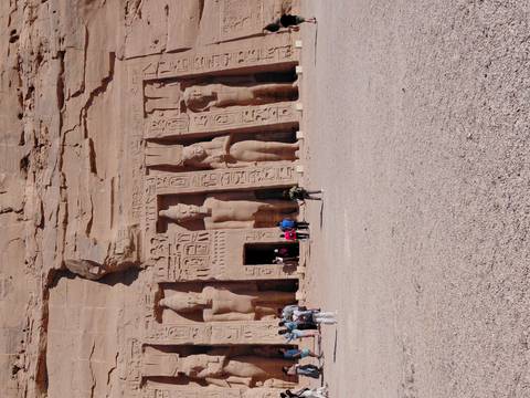       Carved statues on the facade of the Small Temple at Abu Simbel with tourists approaching across open ground
  