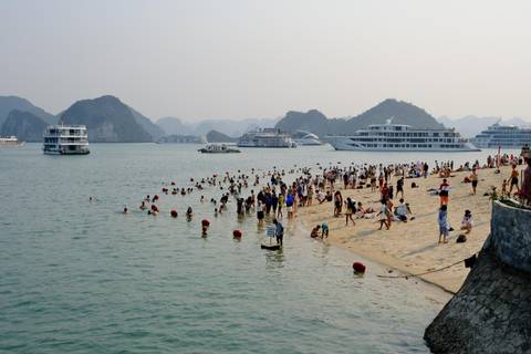       Crowds of swimmers enjoy a sandy beach while cruise ships anchor among the limestone karsts of Halong Bay.
  
