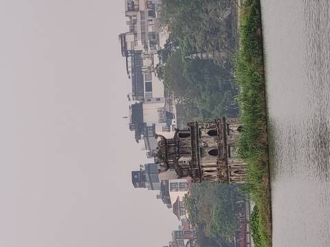       The historic Turtle Tower stands on a small grassy islet in Hoan Kiem Lake with Hanoi's skyline behind on a hazy day.
  