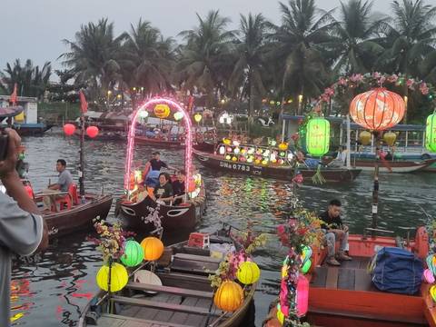       Colorful lantern-lit boats parade on the river during Hoi An's evening festival with palm trees silhouetted behind.
  