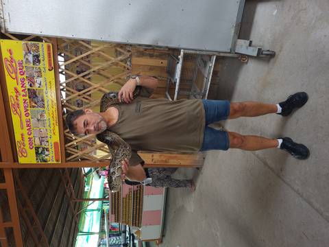       A tourist poses with a large python draped over his shoulders inside a rustic market building.
  