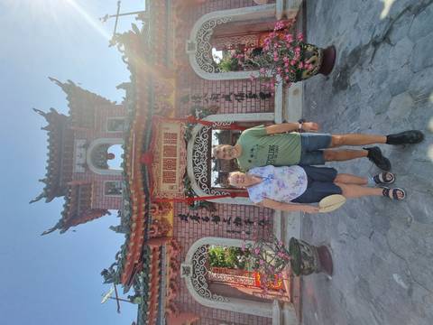       Smiling couple stand before the ornate gateway of the Phuoc Kien Assembly Hall in bright sunshine.
  