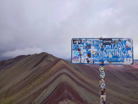       A sticker-covered altitude sign for Vinicunca stands before the striped slopes of Rainbow Mountain under moody clouds.
  