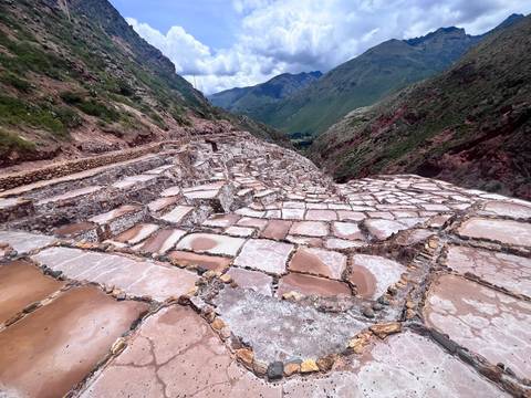       Close-up perspective of the geometric pink-hued evaporation ponds at the Maras salt mines against green mountains.
  