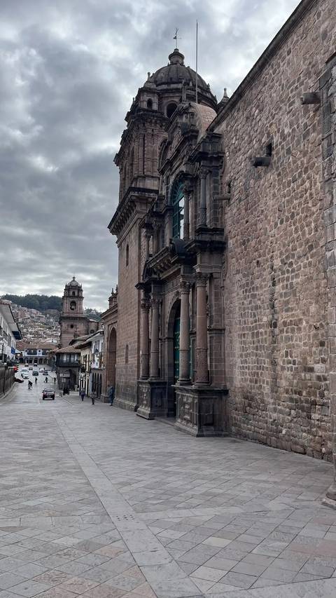       Stone façade of Cusco Cathedral lines a narrow street with the bell tower of Iglesia de la Compañía visible under a grey sky.
  