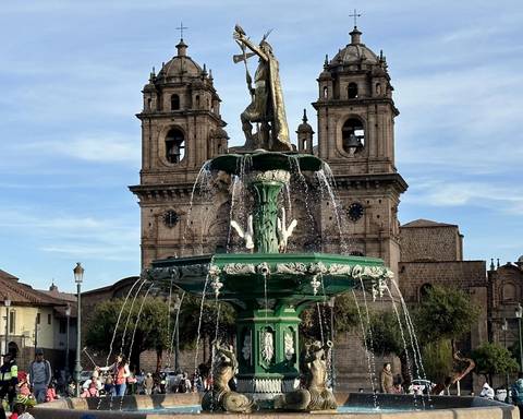      Ornate green fountain topped with an Incan figure splashes before the twin bell towers of Cusco's Iglesia La Compañía.
  