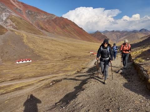       Trekkers with poles stride along a high-altitude valley flanked by ochre and red mountains near Rainbow Mountain.
  