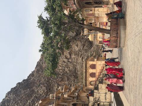       Visitors in colorful sarees walk through a stone courtyard beneath a steep rocky cliff and historic temple buildings.
  