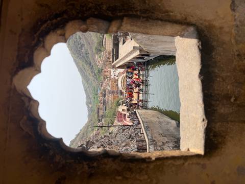       View through an ornate arched window to a sacred pool filled with pilgrims and surrounded by hills.
  