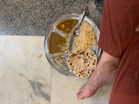       Simple Indian meal of dal, rice and chapati served on a steel tray on the floor with a person’s bare foot visible.
  