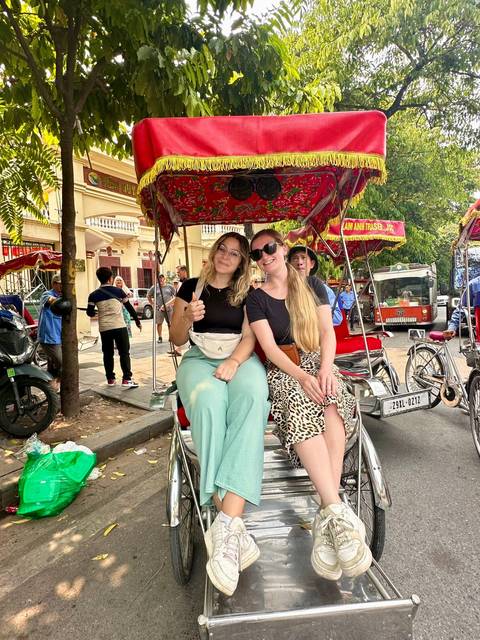       Two women seated on a bright rickshaw enjoying a ride through a Vietnamese city street
  