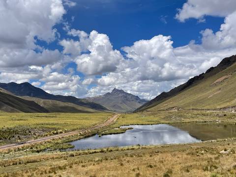       Wide Andean valley with railroad tracks, reflective pond and distant jagged peak beneath billowing clouds
  