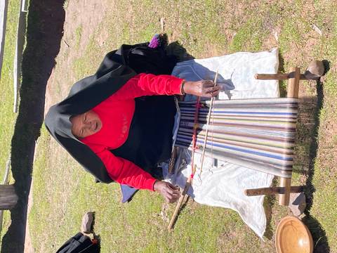       Indigenous woman weaving colorful textiles on a backstrap loom while kneeling on the grass
  